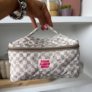 May include: A beige and white checkered quilted cosmetic bag with a brown zipper and handle. The bag has a pink label that reads "BOHO RANCH SHOP". The bag is held by a person wearing a silver bracelet.