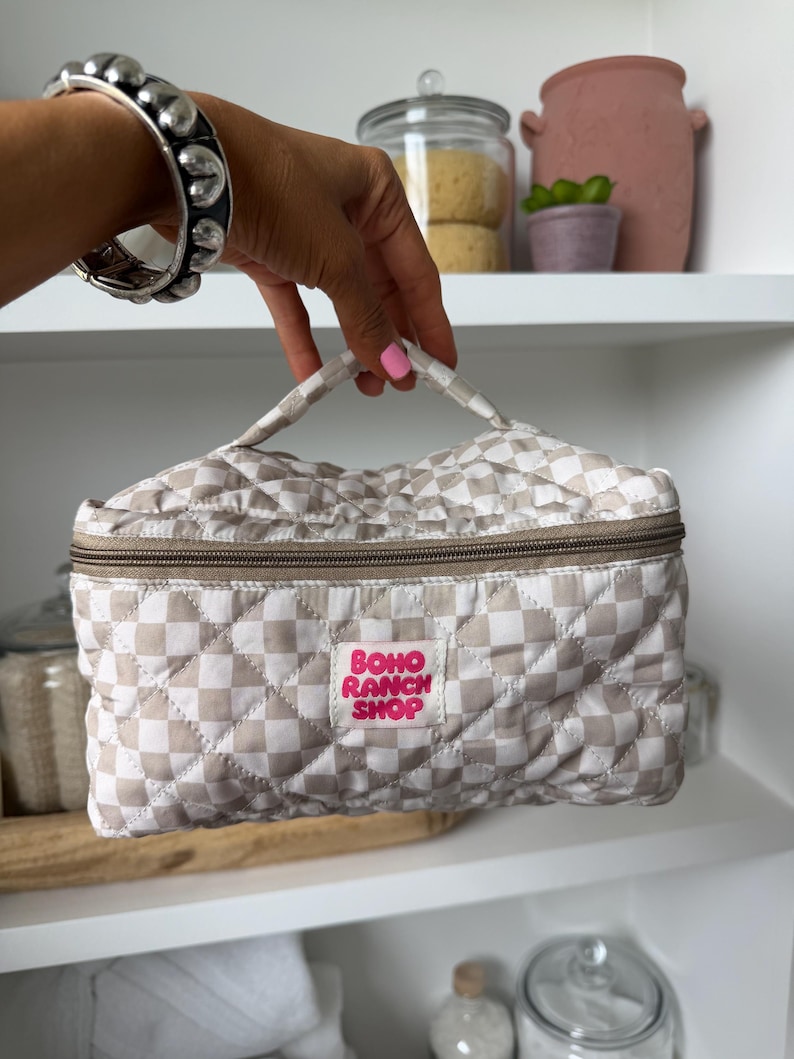 May include: A beige and white checkered quilted cosmetic bag with a brown zipper and handle. The bag has a pink label that reads "BOHO RANCH SHOP". The bag is held by a person wearing a silver bracelet.