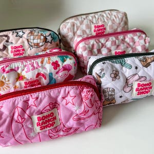 May include: A collection of six quilted zippered pouches in various colors and patterns, including pink, white, and checkered designs. Each pouch features a rectangular fabric label with the text Boho Ranch Shop printed in red letters.