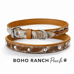 May include: Two dog collars. The top collar is light brown leather with a silver buckle and a section of cowhide. The bottom collar is brown cowhide with silver studs. The text "BOHO RANCH Pooch" is at the bottom.