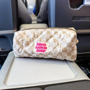 May include: A rectangular quilted pouch featuring a beige and white checkered pattern. A white fabric label is stitched to the front with the text BOHO RANCH SHOP printed in bright pink letters. The pouch has a brown zipper closure along the top.