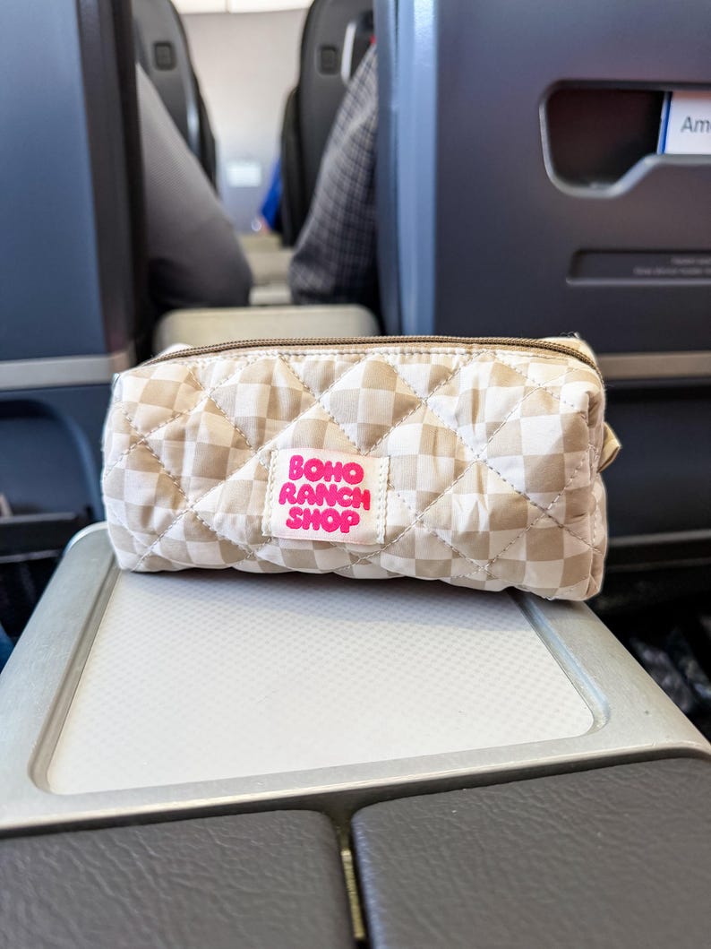 May include: A rectangular quilted pouch featuring a beige and white checkered pattern. A white fabric label is stitched to the front with the text BOHO RANCH SHOP printed in bright pink letters. The pouch has a brown zipper closure along the top.