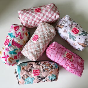 May include: Six quilted fabric pouches in various patterns including pink checkered, western motifs with cowboy boots and cacti, and disco balls. Each rectangular pouch features a white fabric label with the text Boho Ranch Shop in red.