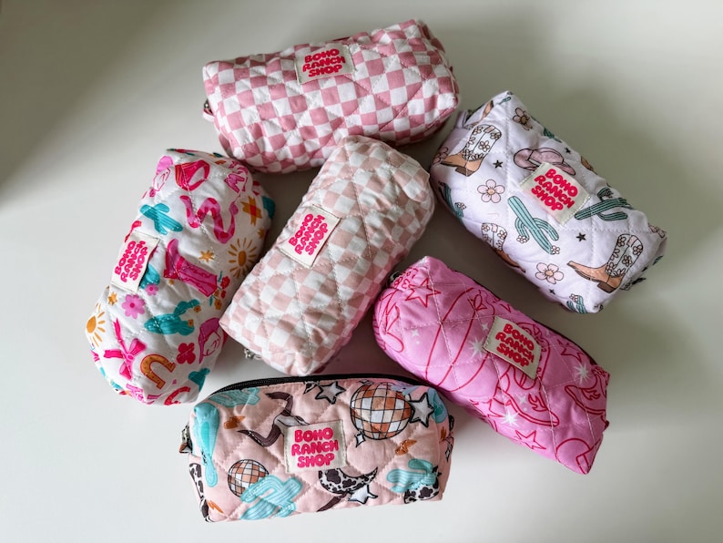 May include: Six quilted fabric pouches in various patterns including pink checkered, western motifs with cowboy boots and cacti, and disco balls. Each rectangular pouch features a white fabric label with the text Boho Ranch Shop in red.