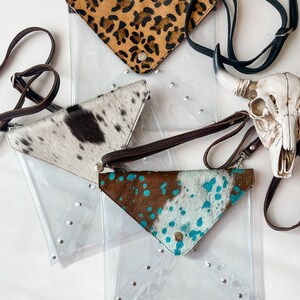 May include: Three clear plastic envelope-style crossbody bags with triangular flaps featuring animal print cowhide textures in leopard, black and white, and brown and turquoise. Each bag has a metal snap closure, silver studs, and a leather strap.