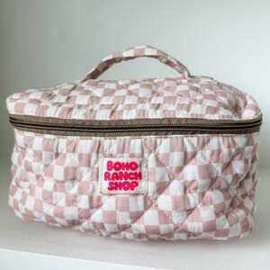 May include: A quilted cosmetic bag with a pink and white checkered pattern. The bag has a brown zipper and a handle. The label reads "BOHO RANCH SHOP".