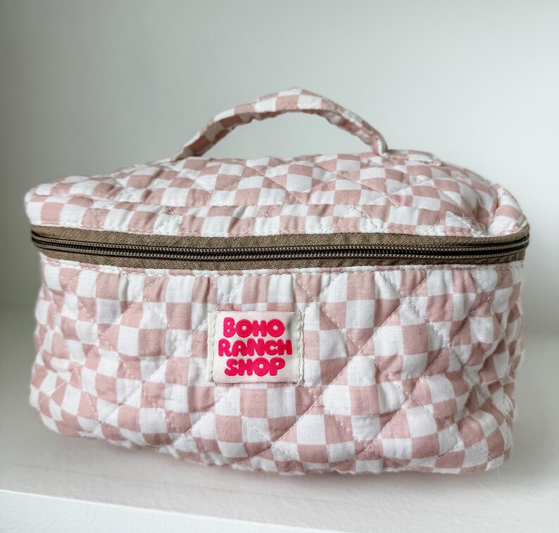 May include: A quilted cosmetic bag with a pink and white checkered pattern. The bag has a brown zipper and a handle. The label reads "BOHO RANCH SHOP".