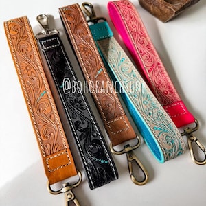 May include: A collection of leather wrist straps in various colors, including tan, black, brown, teal, and pink. Each strap features a floral embossed design and a metal clasp. The straps are approximately 8 inches long.