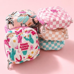 May include: A stack of small, quilted pouches in various patterns and colors. Designs include a pink and white checkerboard, cacti, cowboy boots, and the text "BOHO RANCH SHOP". Each pouch has a zipper closure and a small handle.