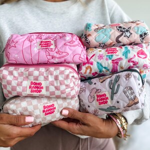 May include: A collection of five quilted rectangular pouches featuring various patterns including pink cowboy boots, checkered prints, cacti, and flowers. Each pouch has a white fabric label with the text Boho Ranch Shop in pink letters.