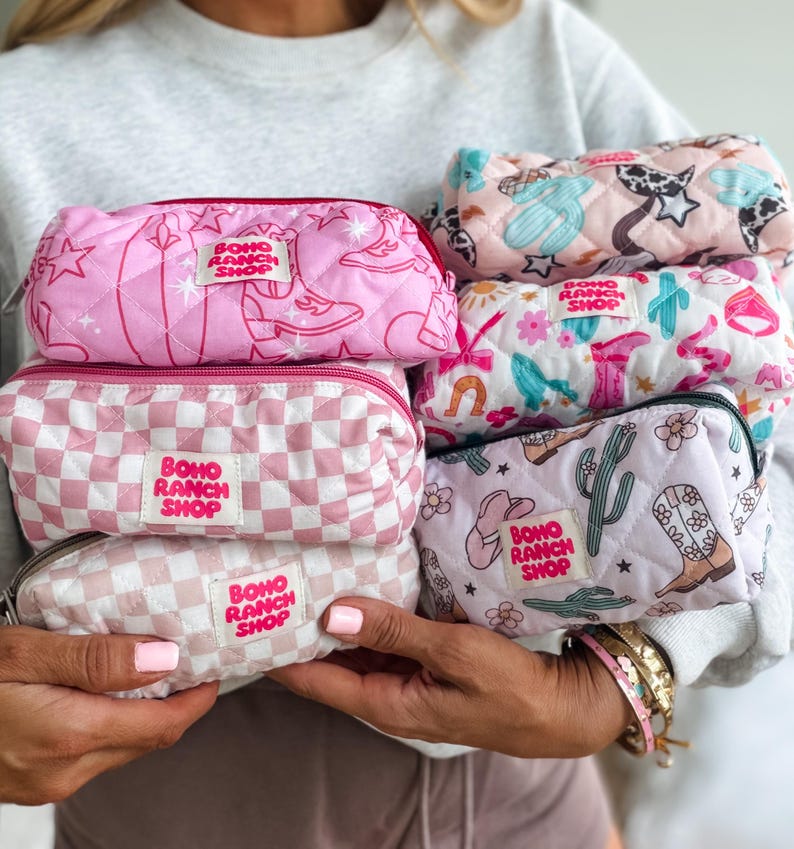 May include: A collection of five quilted rectangular pouches featuring various patterns including pink cowboy boots, checkered prints, cacti, and flowers. Each pouch has a white fabric label with the text Boho Ranch Shop in pink letters.