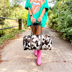 May include: A woman wearing a green t-shirt with the Rolling Stones logo and pink cowboy boots is carrying a brown and white patchwork leather duffel bag.