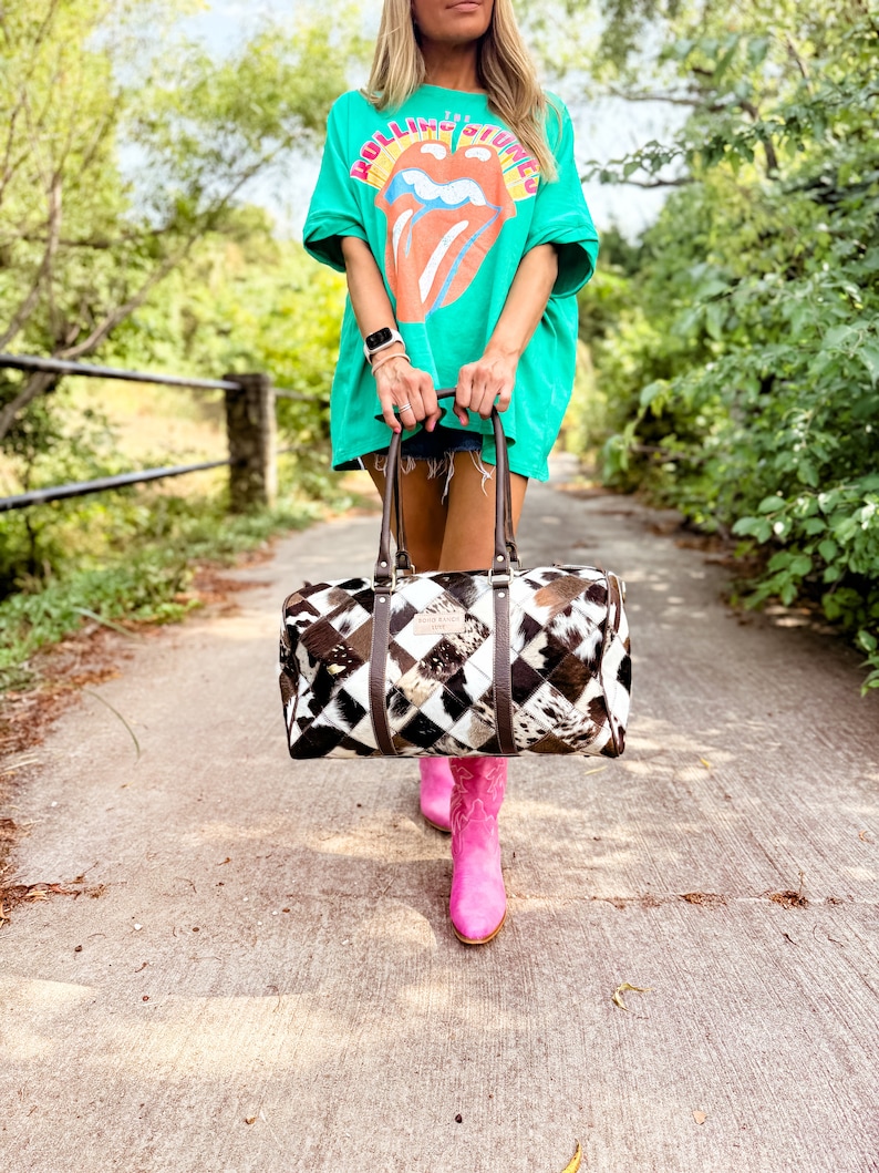 May include: A woman wearing a green t-shirt with the Rolling Stones logo and pink cowboy boots is carrying a brown and white patchwork leather duffel bag.