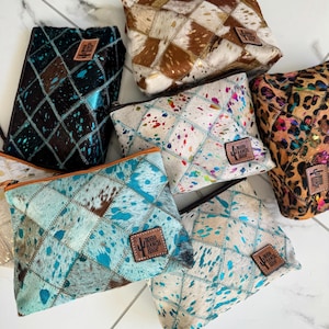 May include: Six quilted cosmetic bags with different patterns. The bags are made of cowhide and have a turquoise and brown color scheme. Each bag has a brown leather patch with the text "Round Ranch Shop" on it.