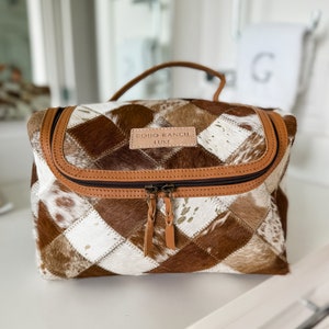 May include: A rectangular toiletry bag featuring a patchwork design of brown and white cowhide squares. The bag has a tan leather handle, tan leather trim, and a zippered closure with two braided leather pulls. A tan leather label reads BOHO RANCH LUXE.