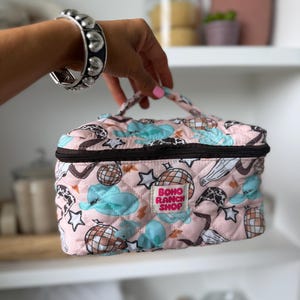 May include: A pink quilted cosmetic bag with a black zipper and handle. The bag features a pattern of disco balls, stars, and clouds in blue and brown. The text "BOHO RANCH SHOP" is printed on the front.
