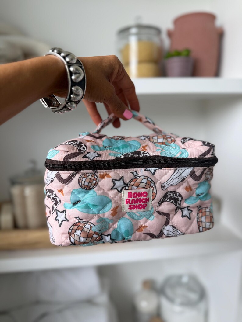 May include: A pink quilted cosmetic bag with a black zipper and handle. The bag features a pattern of disco balls, stars, and clouds in blue and brown. The text "BOHO RANCH SHOP" is printed on the front.