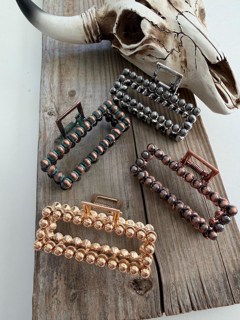 May include: Four rectangular hair claw clips in various metallic finishes: gold, copper, bronze, and silver. Each clip is adorned with a series of small, round beads. The clips are arranged on a weathered wooden surface, with a cow skull in the background.