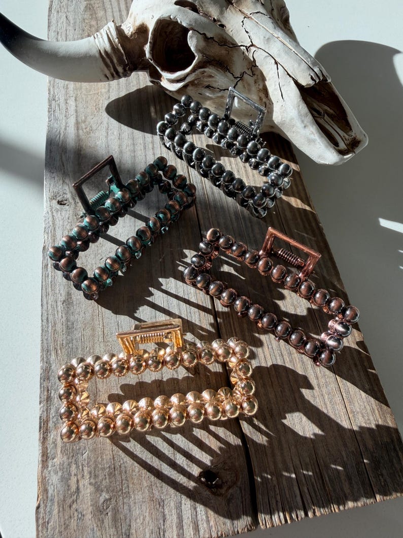 May include: Four beaded hair claw clips in various metallic finishes: gold, copper, bronze, and gunmetal. The clips are rectangular and feature a series of small, round beads. The background includes a weathered wooden surface and a cow skull.