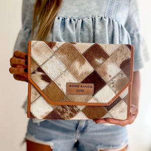 May include: A brown and white patchwork leather clutch with a brown leather flap and a gold label that reads "BOHO RANCH LUXE".