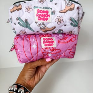 May include: Two quilted zippered pouches stacked vertically. The top pouch is white with a pattern of brown cowboy boots, green cacti, and pink flowers. The bottom pouch is pink with a red line-art pattern of boots and stars. Both feature a white label with the text BOHO RANCH SHOP in pink.