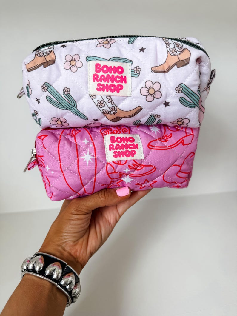 May include: Two quilted zippered pouches stacked vertically. The top pouch is white with a pattern of brown cowboy boots, green cacti, and pink flowers. The bottom pouch is pink with a red line-art pattern of boots and stars. Both feature a white label with the text BOHO RANCH SHOP in pink.