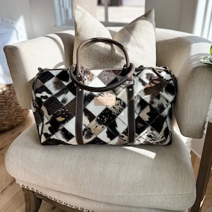May include: A brown leather and cowhide patchwork travel bag with a brown leather handle and a white label that reads "BOHO RANCH LUXE".