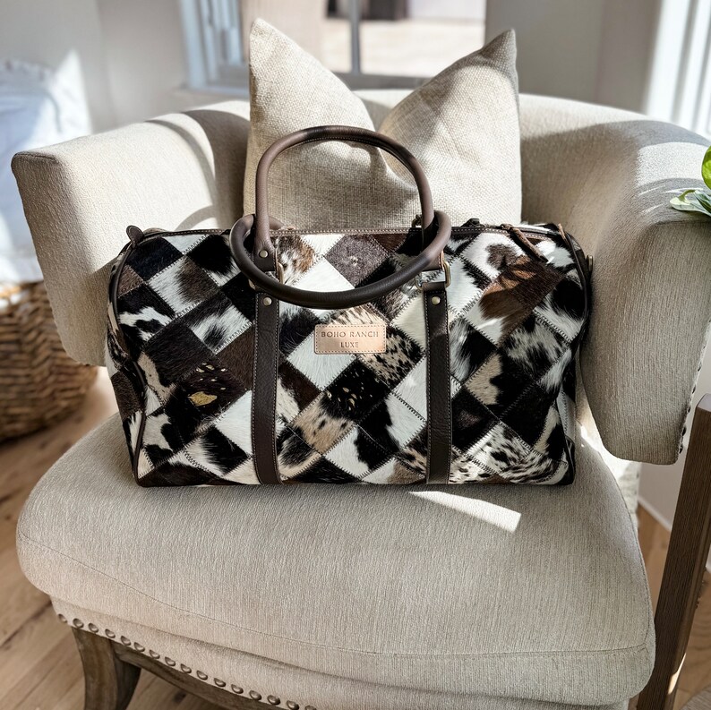 May include: A brown leather and cowhide patchwork travel bag with a brown leather handle and a white label that reads "BOHO RANCH LUXE".