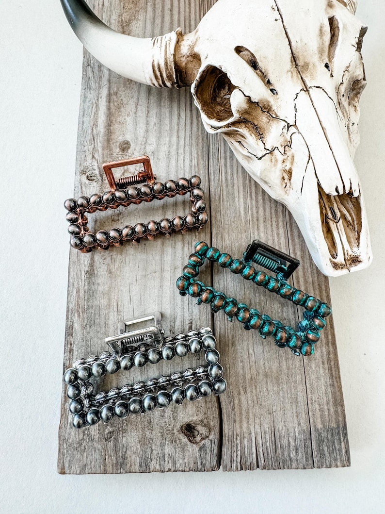 May include: Three rectangular hair claw clips in copper, silver, and teal. Each clip is adorned with small, round beads. The clips are arranged on a weathered wooden surface, with a cow skull in the background.