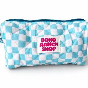 May include: A quilted rectangular zippered pouch featuring a light blue and white checkered pattern. A white rectangular fabric label is sewn onto the center front with the text BOHO RANCH SHOP printed in bold pink letters. The zipper is teal.