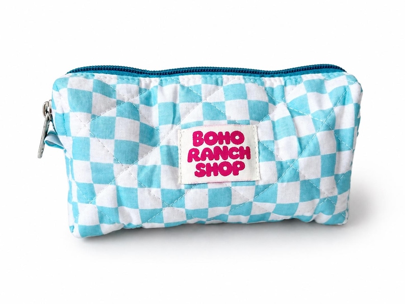 May include: A quilted rectangular zippered pouch featuring a light blue and white checkered pattern. A white rectangular fabric label is sewn onto the center front with the text BOHO RANCH SHOP printed in bold pink letters. The zipper is teal.