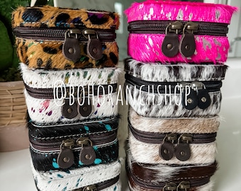 May include: Stack of six small, square, zippered boxes with different cowhide patterns. The boxes are made of leather and have a brown zipper and two brown leather pulls. The boxes are stacked in two columns of three. The boxes are all different colors and patterns. The top box in the left column is leopard print, the middle box is white with colorful paint splatters, and the bottom box is white with blue paint splatters. The top box in the right column is hot pink with silver glitter, the middle box is black and white cowhide, and the bottom box is brown and white cowhide.