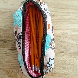 May include: A quilted pencil case with a light pink exterior featuring a pattern of disco balls and stars in blue, brown, and white. The interior is lined with a pink and white gingham fabric and contains several yellow wooden pencils.