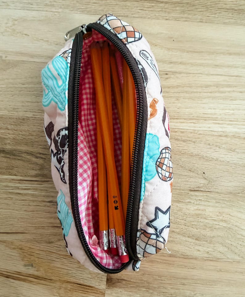 May include: A quilted pencil case with a light pink exterior featuring a pattern of disco balls and stars in blue, brown, and white. The interior is lined with a pink and white gingham fabric and contains several yellow wooden pencils.