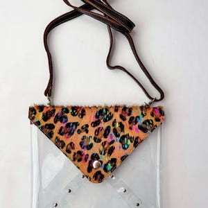 May include: A clear plastic envelope-style crossbody bag featuring a triangular flap made of tan animal-print hair-on hide with iridescent accents. The bag includes a brown leather strap and silver-toned metal hardware, including a snap closure.