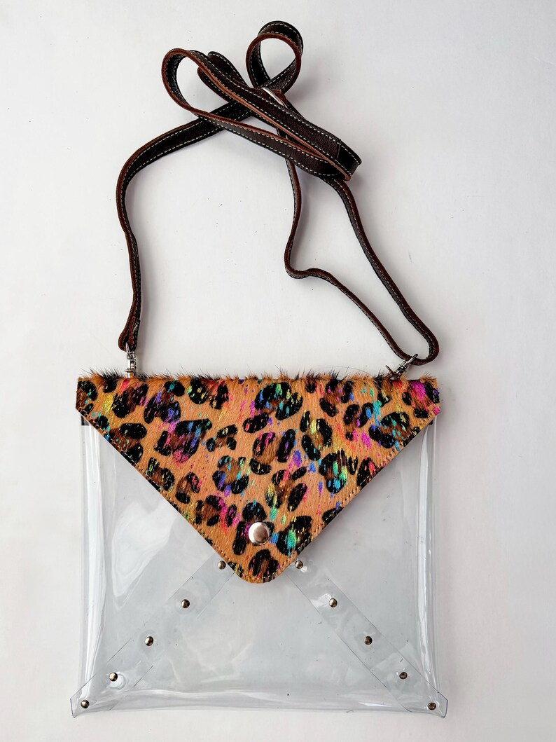 May include: A clear plastic envelope-style crossbody bag featuring a triangular flap made of tan animal-print hair-on hide with iridescent accents. The bag includes a brown leather strap and silver-toned metal hardware, including a snap closure.