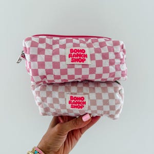 May include: Two stacked rectangular quilted pouches featuring a pink and white checkered pattern. Each pouch has a central white fabric label with the text BOHO RANCH SHOP printed in bold pink letters. The pouches include a pink zipper closure.