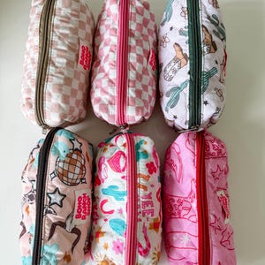 May include: Six rectangular quilted pouches with zippered closures. Designs include pink and white checkered patterns, cactus and cowboy boot prints, disco balls, and western motifs. Each pouch features a small white label reading BOHO RANCH SHOP.
