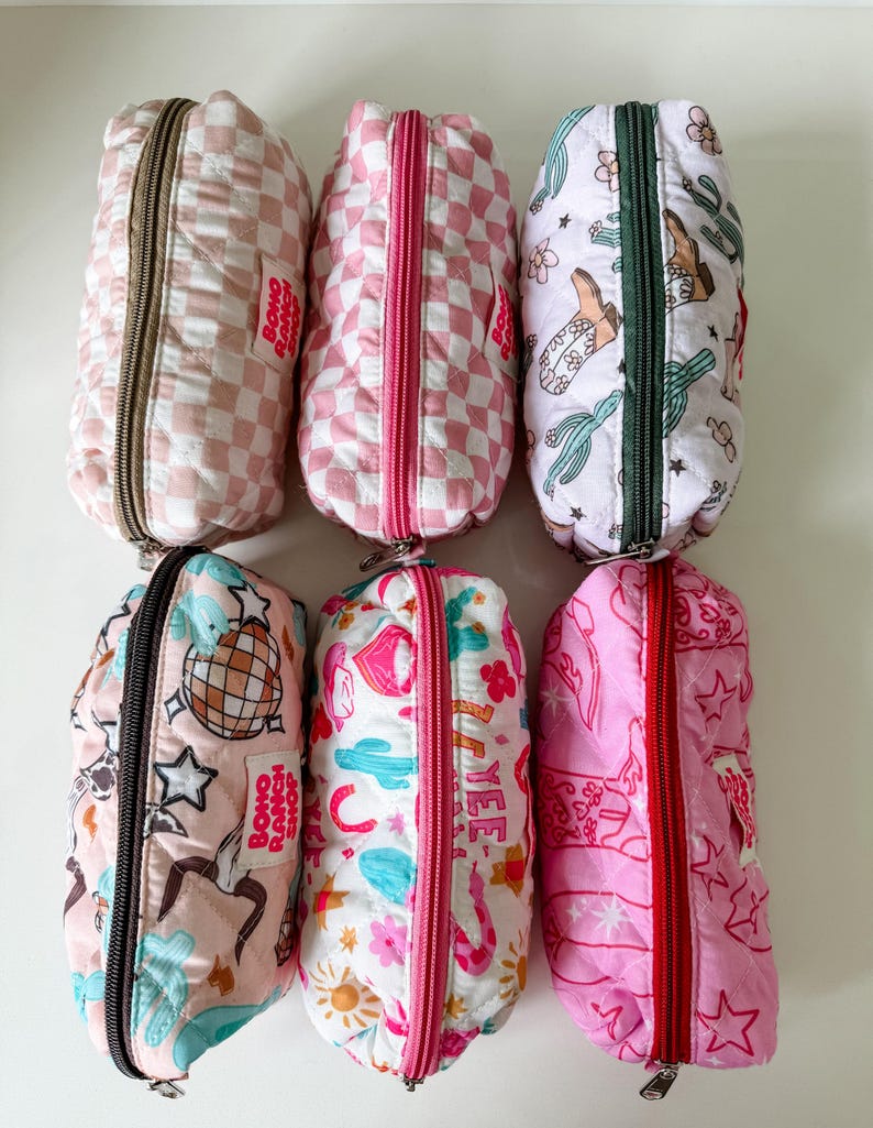 May include: Six rectangular quilted pouches with zippered closures. Designs include pink and white checkered patterns, cactus and cowboy boot prints, disco balls, and western motifs. Each pouch features a small white label reading BOHO RANCH SHOP.