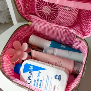 May include: A pink and brown cosmetic bag with a gingham lining, open to reveal various toiletries. The bag contains a bottle of CeraVe Daily Moisturizing Lotion (8 fl oz), a pink flower-shaped hair clip, and other beauty products.