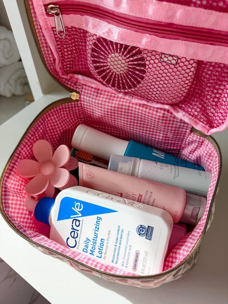 May include: A pink and brown cosmetic bag with a gingham lining, open to reveal various toiletries. The bag contains a bottle of CeraVe Daily Moisturizing Lotion (8 fl oz), a pink flower-shaped hair clip, and other beauty products.