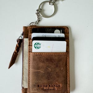 May include: A brown leather wallet with a zipper closure and a key ring. The wallet has a card slot and a small pocket. The wallet is embossed with the text "BOHO RANCH handmade".