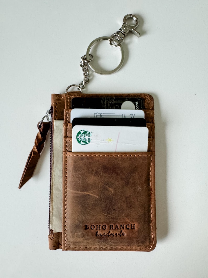 May include: A brown leather wallet with a zipper closure and a key ring. The wallet has a card slot and a small pocket. The wallet is embossed with the text "BOHO RANCH handmade".