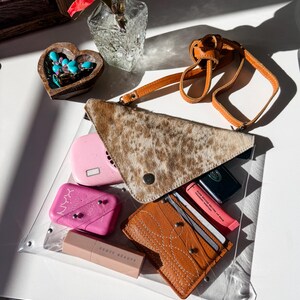 May include: A clear rectangular bag containing a tan and brown cowhide triangular pouch with a strap, a pink NYX blush compact, a Fenty Beauty lipstick, a brown leather card holder with white stitching, and a pink lip balm tube.