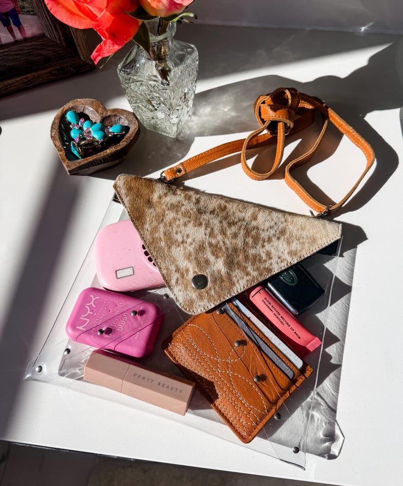 May include: A clear rectangular bag containing a tan and brown cowhide triangular pouch with a strap, a pink NYX blush compact, a Fenty Beauty lipstick, a brown leather card holder with white stitching, and a pink lip balm tube.