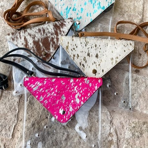 May include: A collection of four clear vinyl crossbody bags featuring triangular cowhide flaps. The flaps come in various patterns including turquoise splatter, brown and white hair-on hide, gold metallic speckle, and hot pink with silver foil.