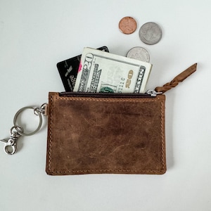 May include: A brown leather zippered pouch with a key ring attached. The pouch is open and contains a credit card and a twenty dollar bill.