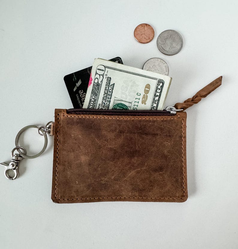 May include: A brown leather zippered pouch with a key ring attached. The pouch is open and contains a credit card and a twenty dollar bill.