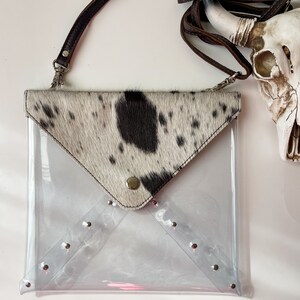 May include: A clear plastic envelope-style crossbody bag featuring a cowhide flap with black and white spots. The bag includes a brown leather strap and silver-toned metal studs along the bottom edges of the transparent body and the flap closure.