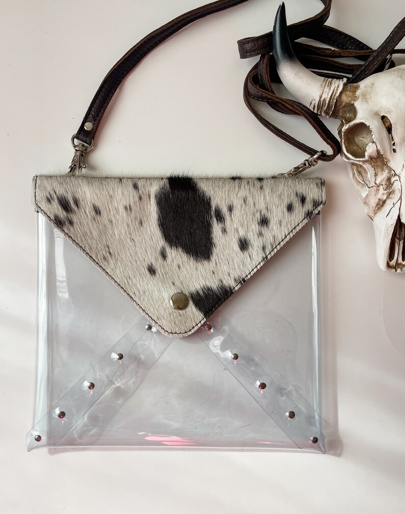 May include: A clear plastic envelope-style crossbody bag featuring a cowhide flap with black and white spots. The bag includes a brown leather strap and silver-toned metal studs along the bottom edges of the transparent body and the flap closure.